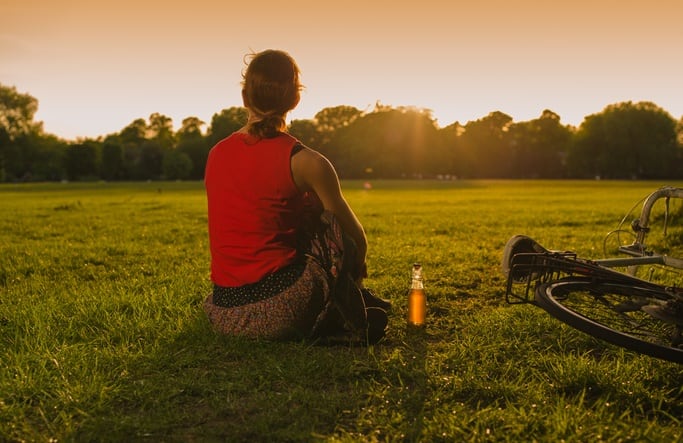 A young woman is admiring the sunset in a park there is a bottle and a bicycle next to her
