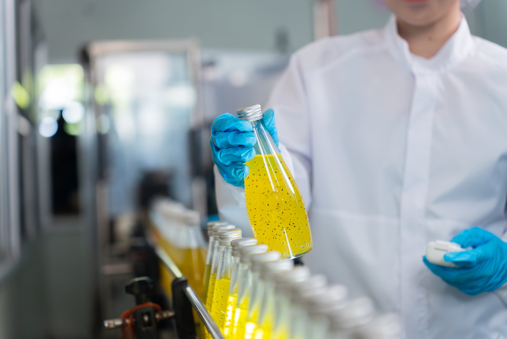 A young adult worker wearing a white lab coat, blue gloves, and a hairnet is inspecting a clear glass bottle filled with a yellow beverage containing seeds on an automated production line in a modern beverage manufacturing facility. The worker is ensuring quality control and safety standards are met during the bottling process, highlighting the importance of hygiene and precision in food and drink production environments.