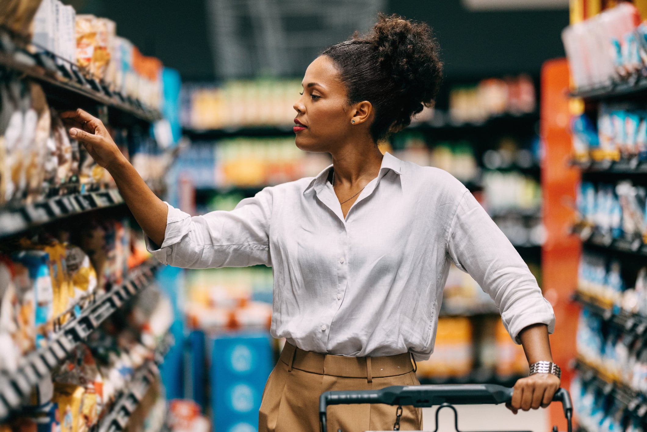 A thoughtful woman shops in a supermarket, browsing through products on the shelves. She seems to be making a careful decision about her purchase.