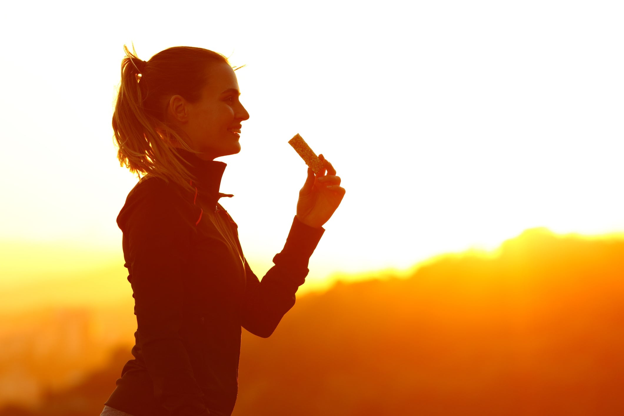 Silhouette of woman eating energy bar after running at sunset in the mountains.