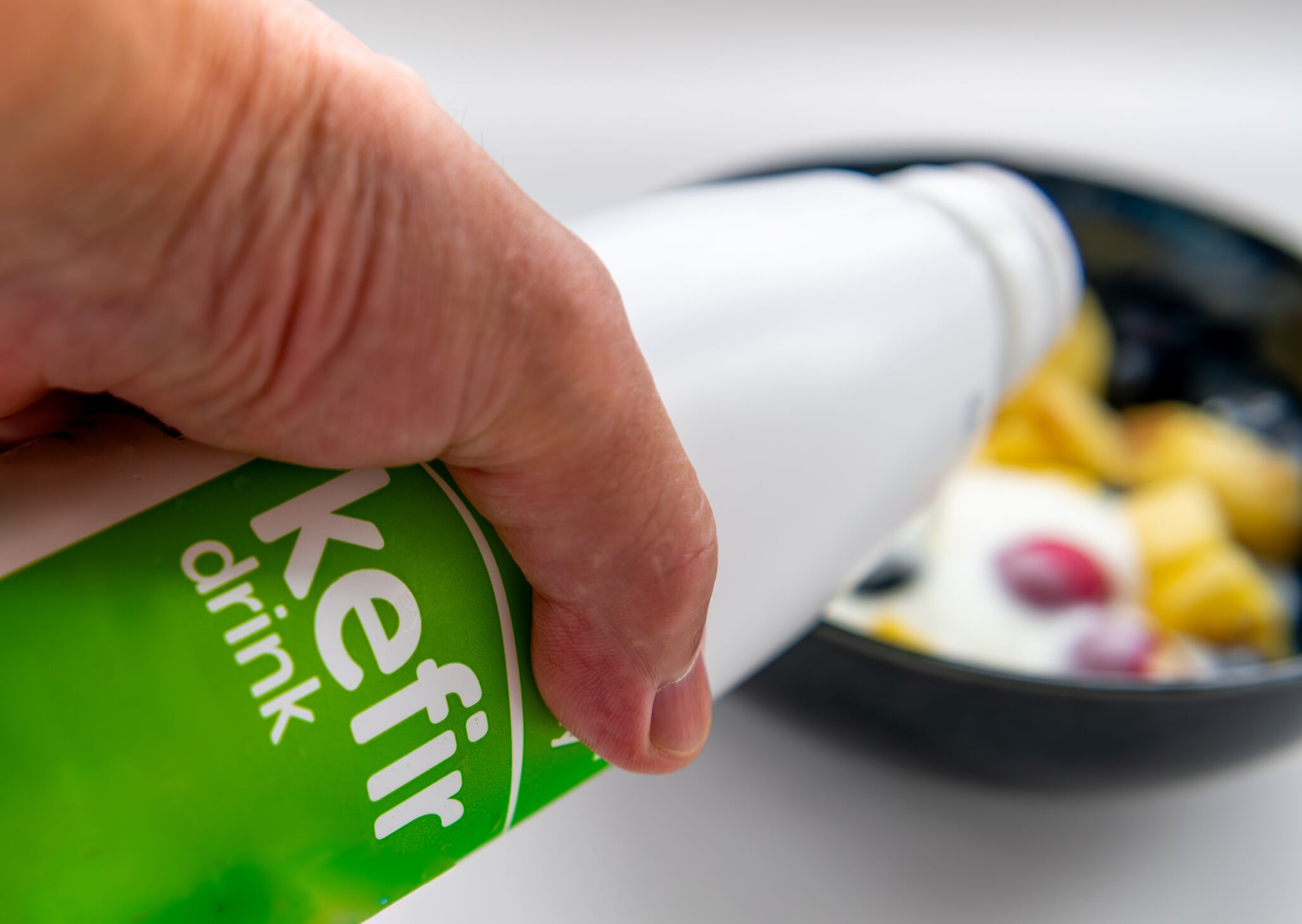 A person pouring Kefir yogurt from a bottle into a bowl of fruit.