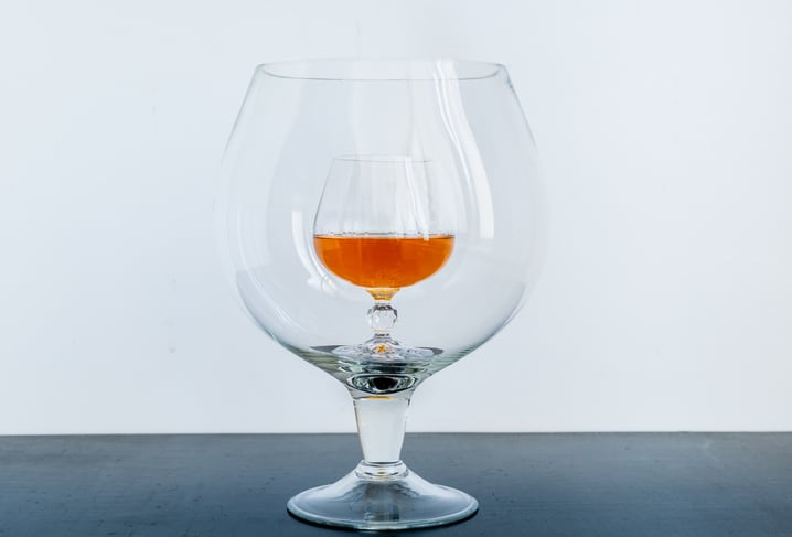 Small glass with whiskey inside big transparent glass on black table on white background