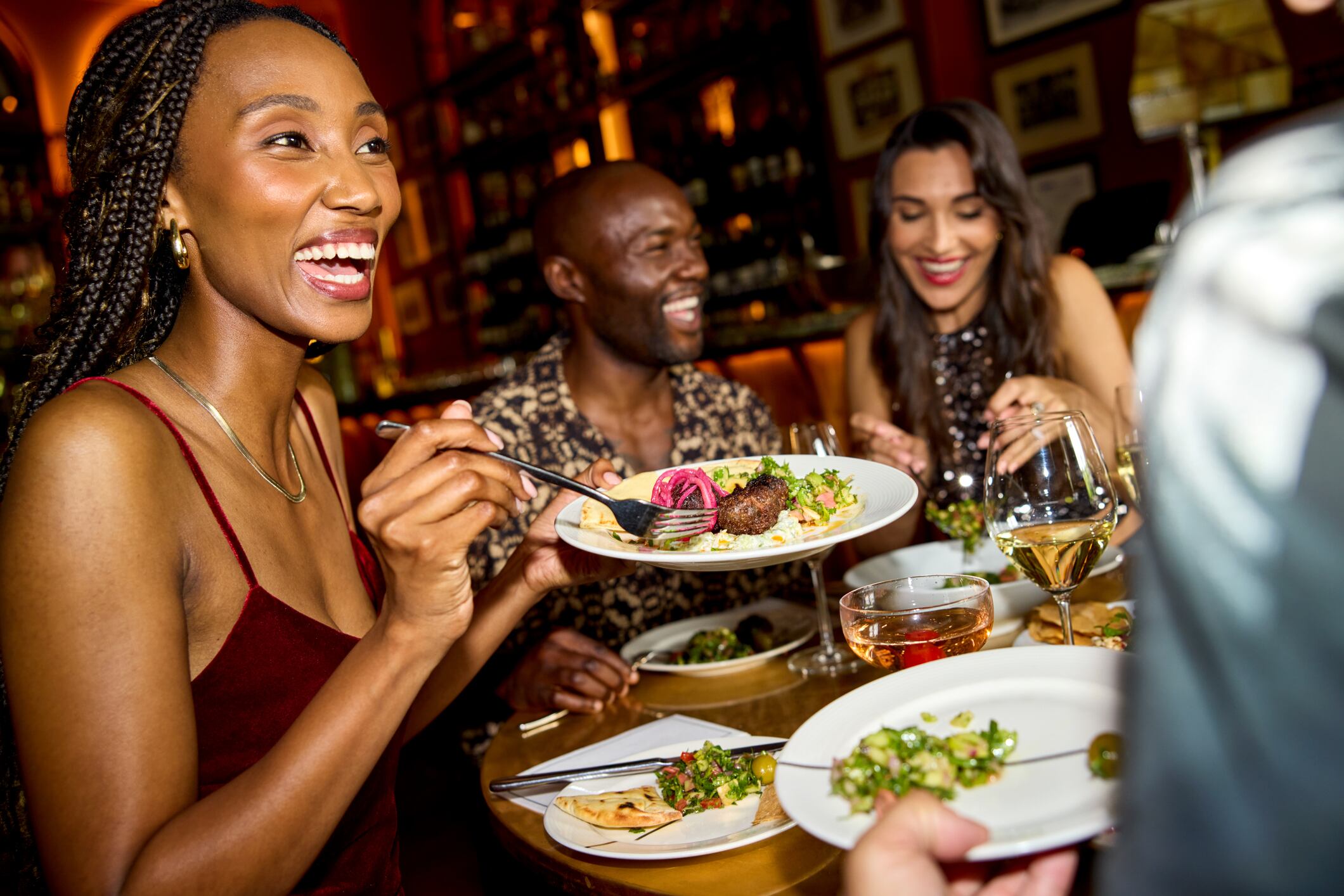 Happy woman having dinner with friends in restaurant. Young female is holding food plate in hand. She is wearing sleeveless dress.