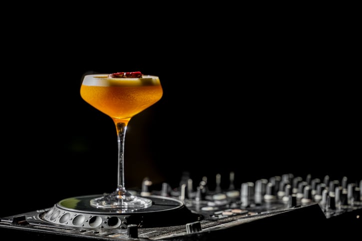 A vibrant orange Porn Star Martini cocktail in a coupe glass with a passion fruit garnish, set on a DJ mixer with dark moody lighting, perfect for nightlife and party themes
