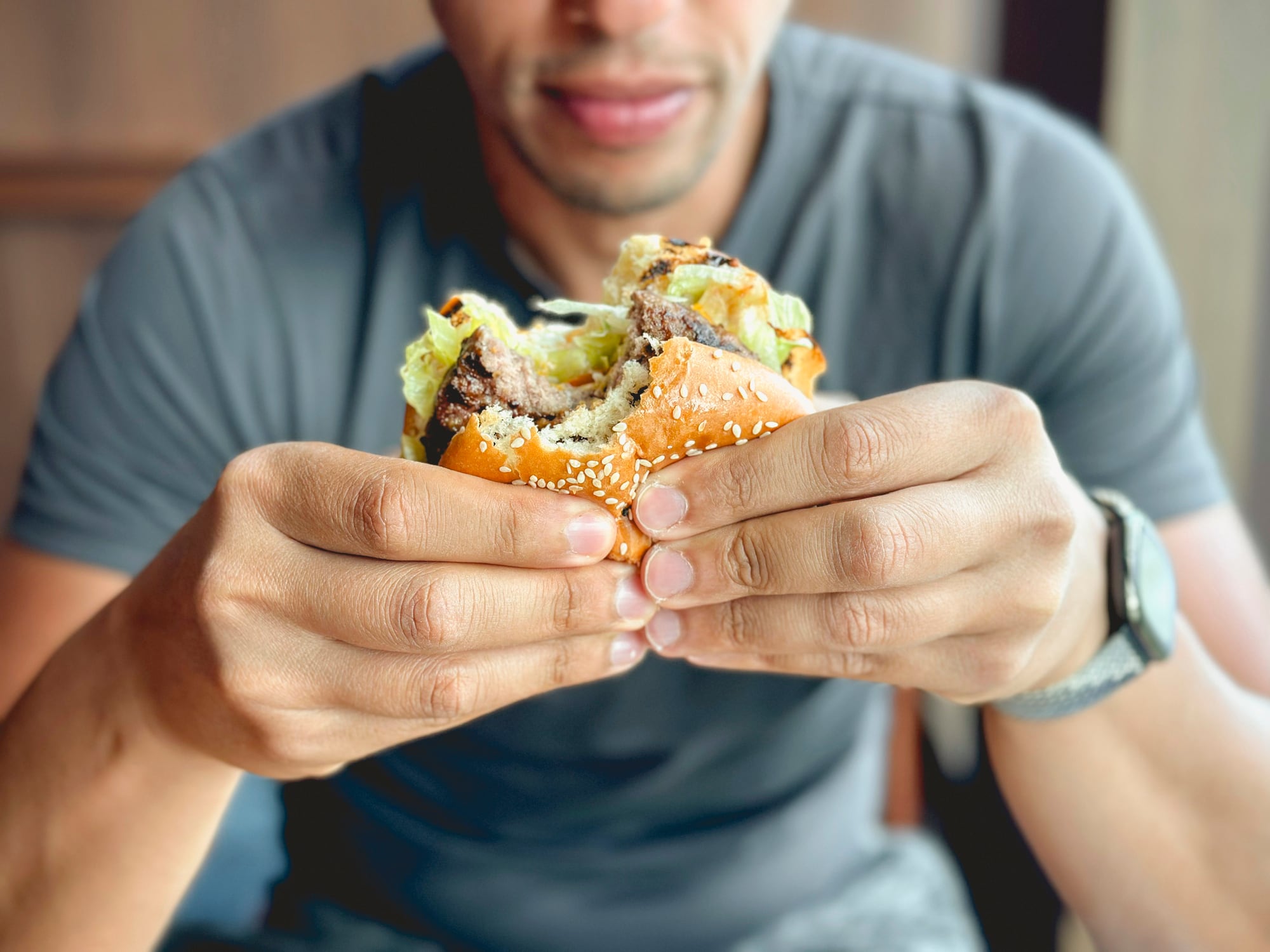 Man in a gray t-shirt prepares to eat a juicy cheeseburger with caramelized onions and lettuce, served with crispy French fries in a metal tray on a wooden table.