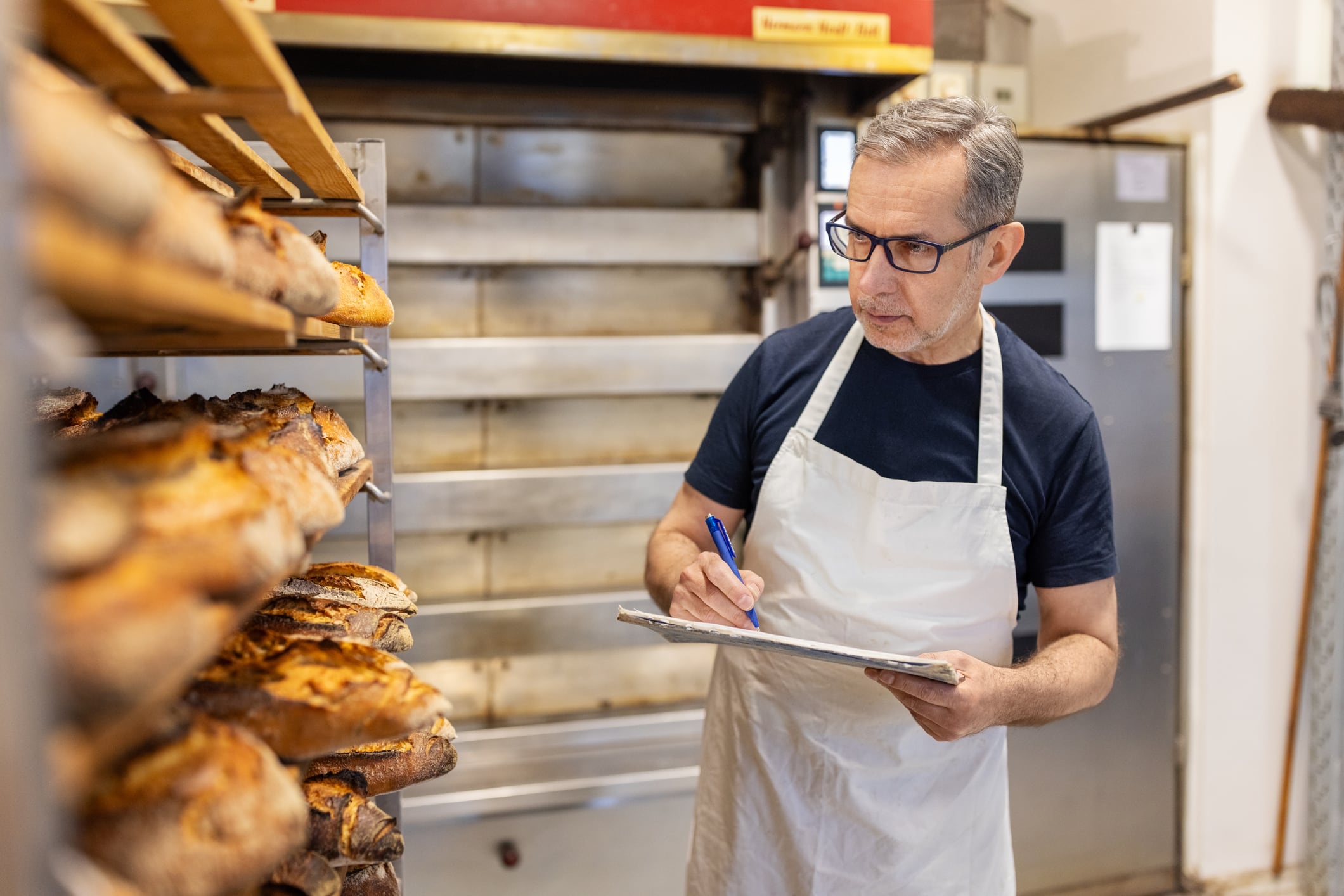 Senior baker carefully checks the freshly baked bread on a rack in the bakery kitchen. Quality control and inventory in traditional bakery kitchen.
