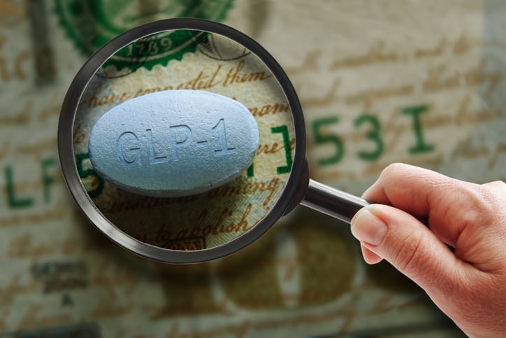 Hand holding a magnifying glass over a GLP-1 pill on a hundred dollar bill