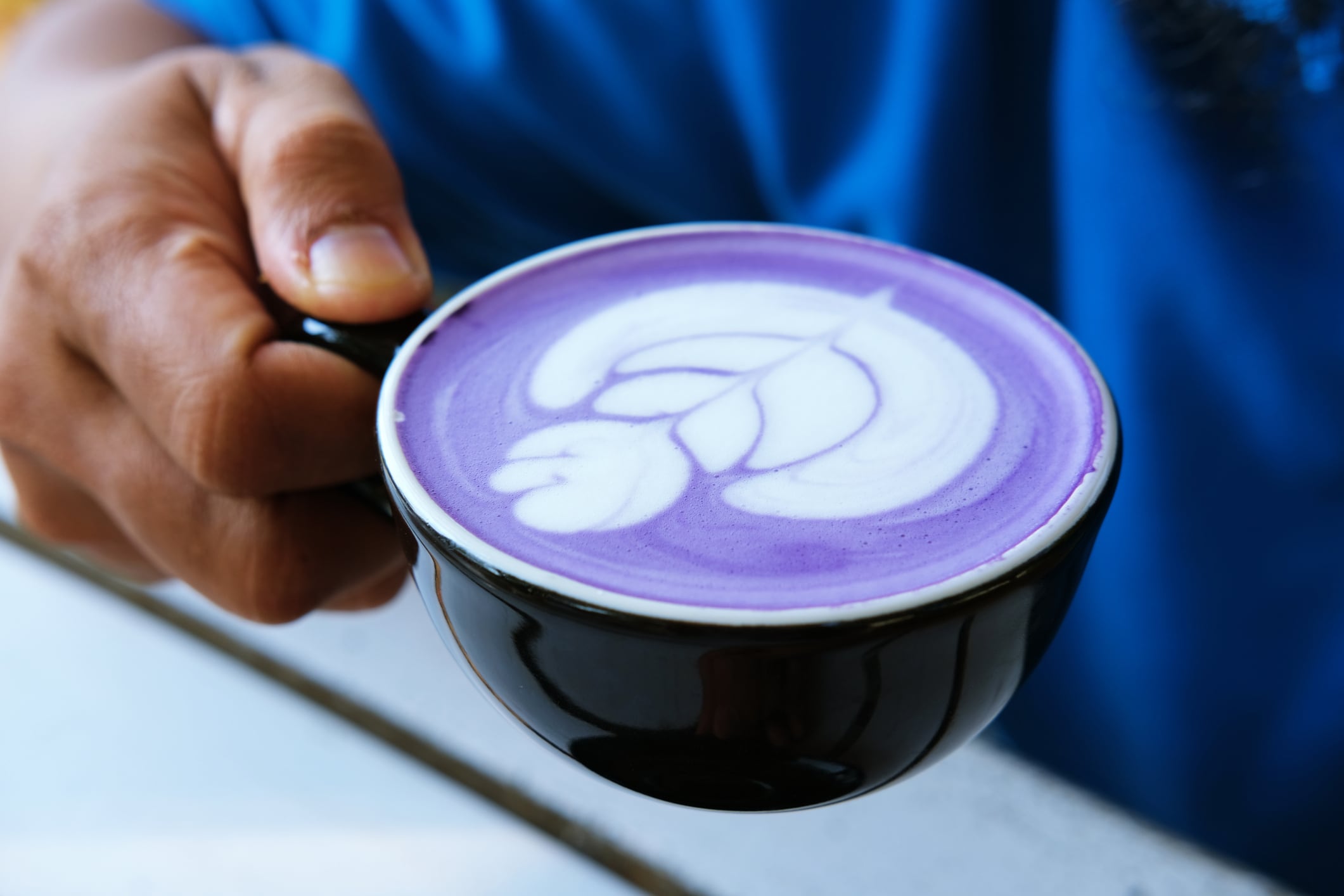Close-up of a hand holding a cup of purple latte art, featuring a detailed leaf design. Perfect for use in cafe promotions, coffee culture blogs, social media, and lifestyle content.