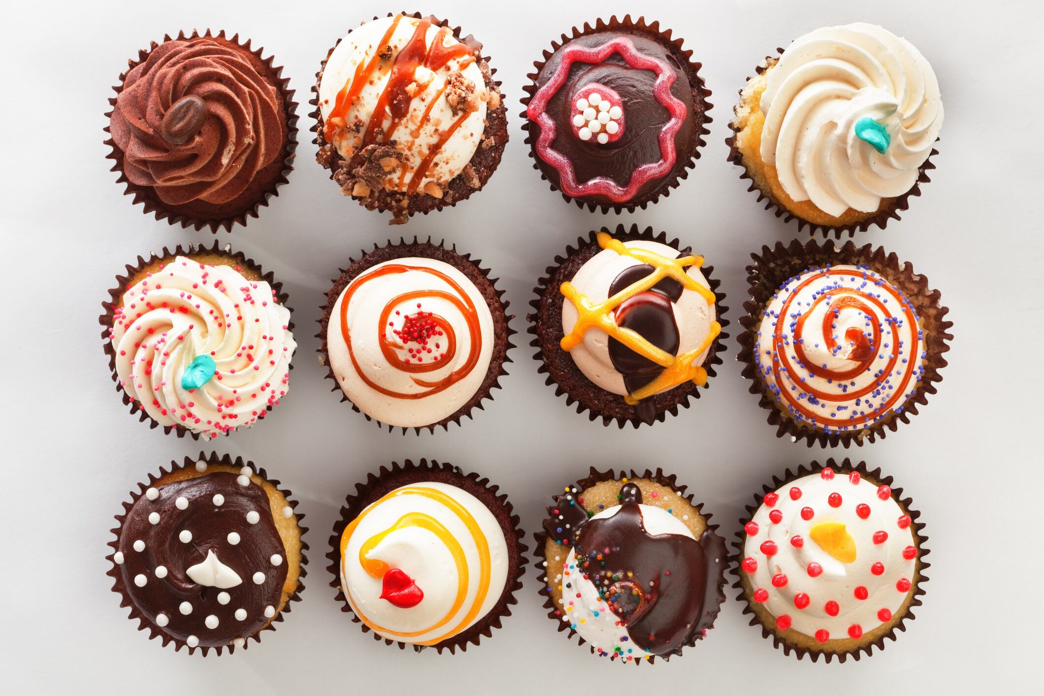 Horizontal high angle, directly overhead view of a dozen frosted cupcakes. The cupcakes are against a white background and are decorated with swirls, sprinkles, drizzles and candies—a tempting decadent treat for those who indulge in sweets and love cakes with lots of toppings.