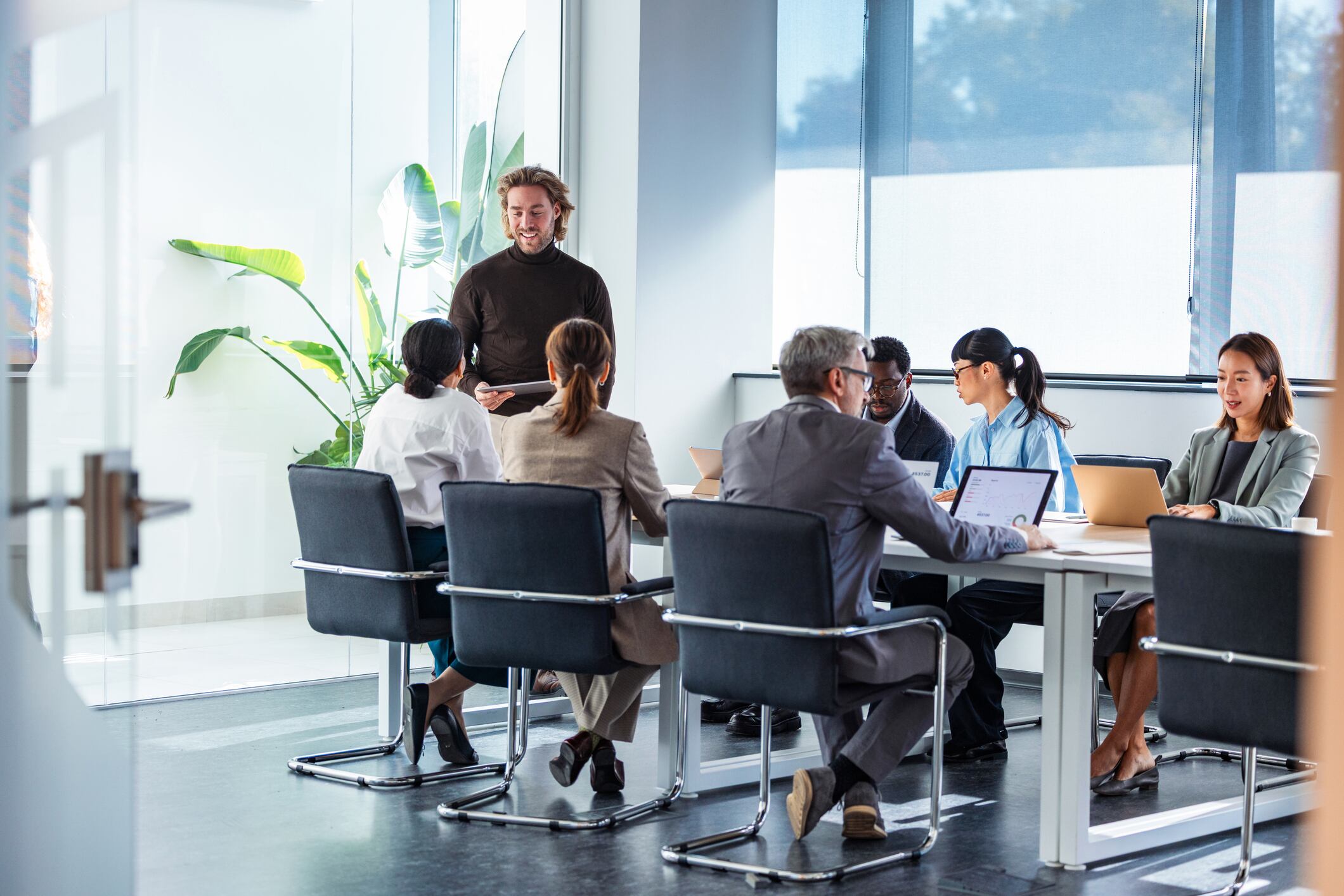 A diverse group of professionals around a conference table engage in a collaborative business meeting. A presenter stands at the head of the table while teammates work on laptops, discussing ideas in a bright, modern office.