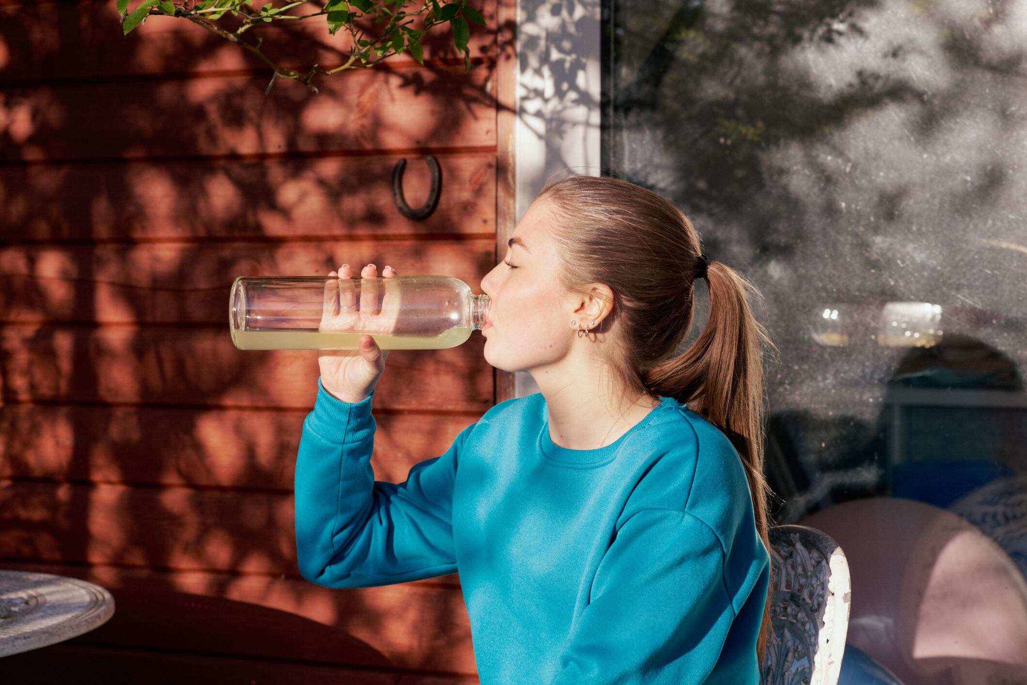UK, Harlow, Essex, young woman sitting drinking water from a reusable bottle after practicing yoga in her garden in springtime.