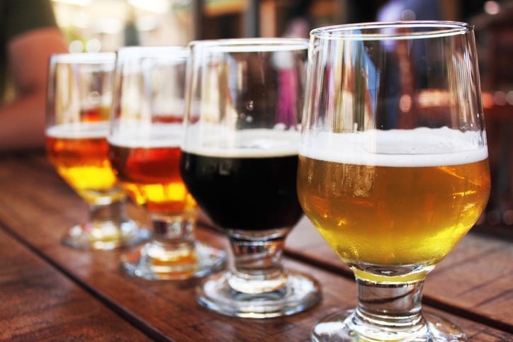 Flight of four beers lined up on a wooden table, with selective focus on the  first beer. Photo taken outdoors with natural lighting; beers range in color from light to very dark. Background out of focus.
