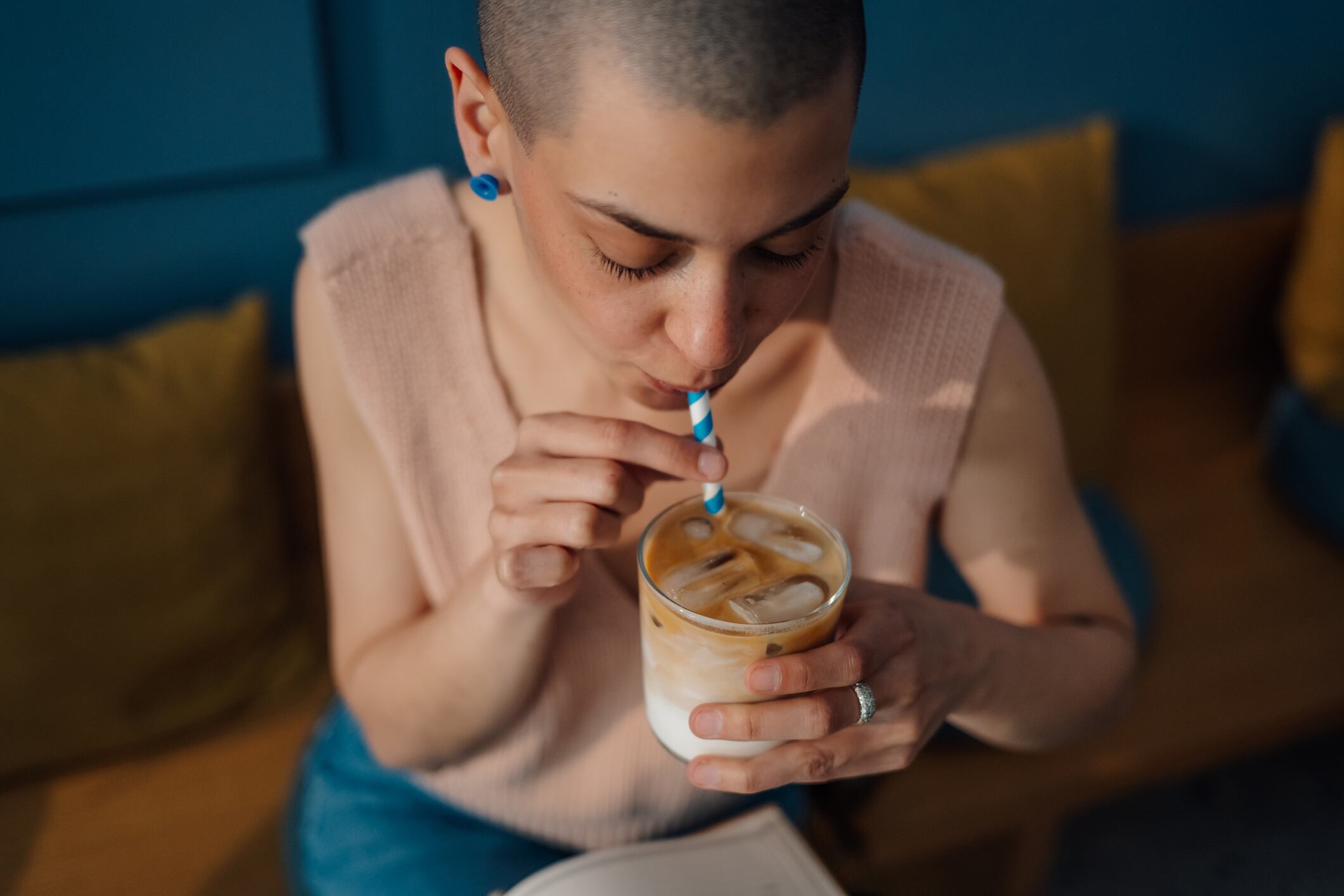 Young woman with a shaved head is sitting in a cafe, savoring an iced coffee through a paper straw