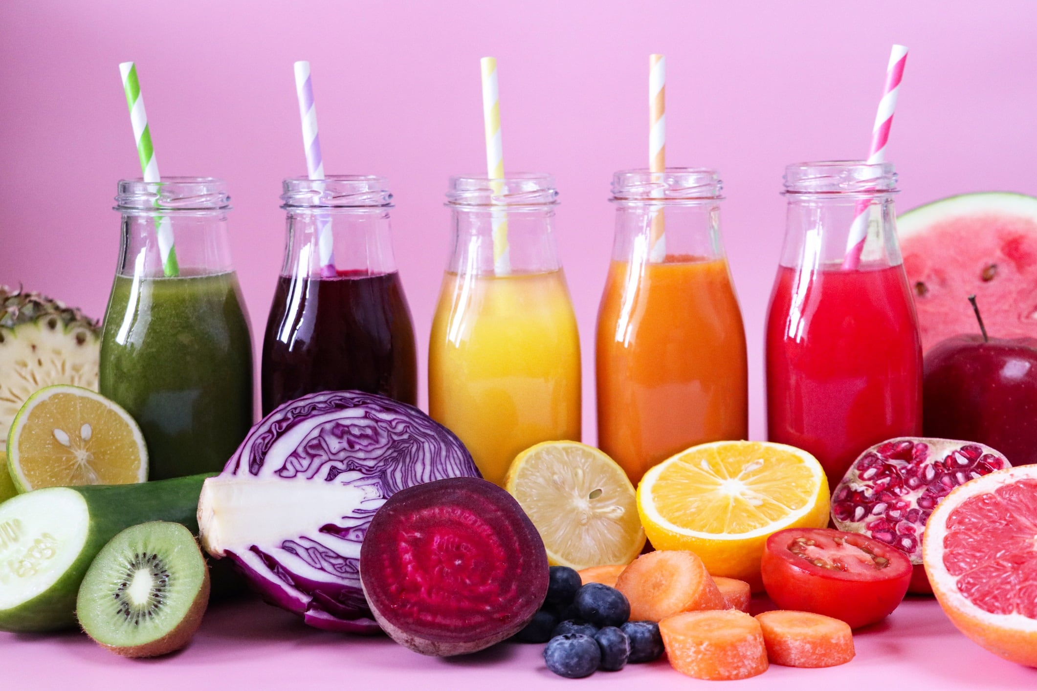 Close-up view of purple, green, yellow, orange and red fruit and vegetable juice smoothies in glass bottles with stripped drinking straws surrounded by watermelon, apple, pomegranate, tomato, carrot, orange, lemon, blueberries, beetroot, red cabbage, kiwi, cucumber, lime and pineapple.