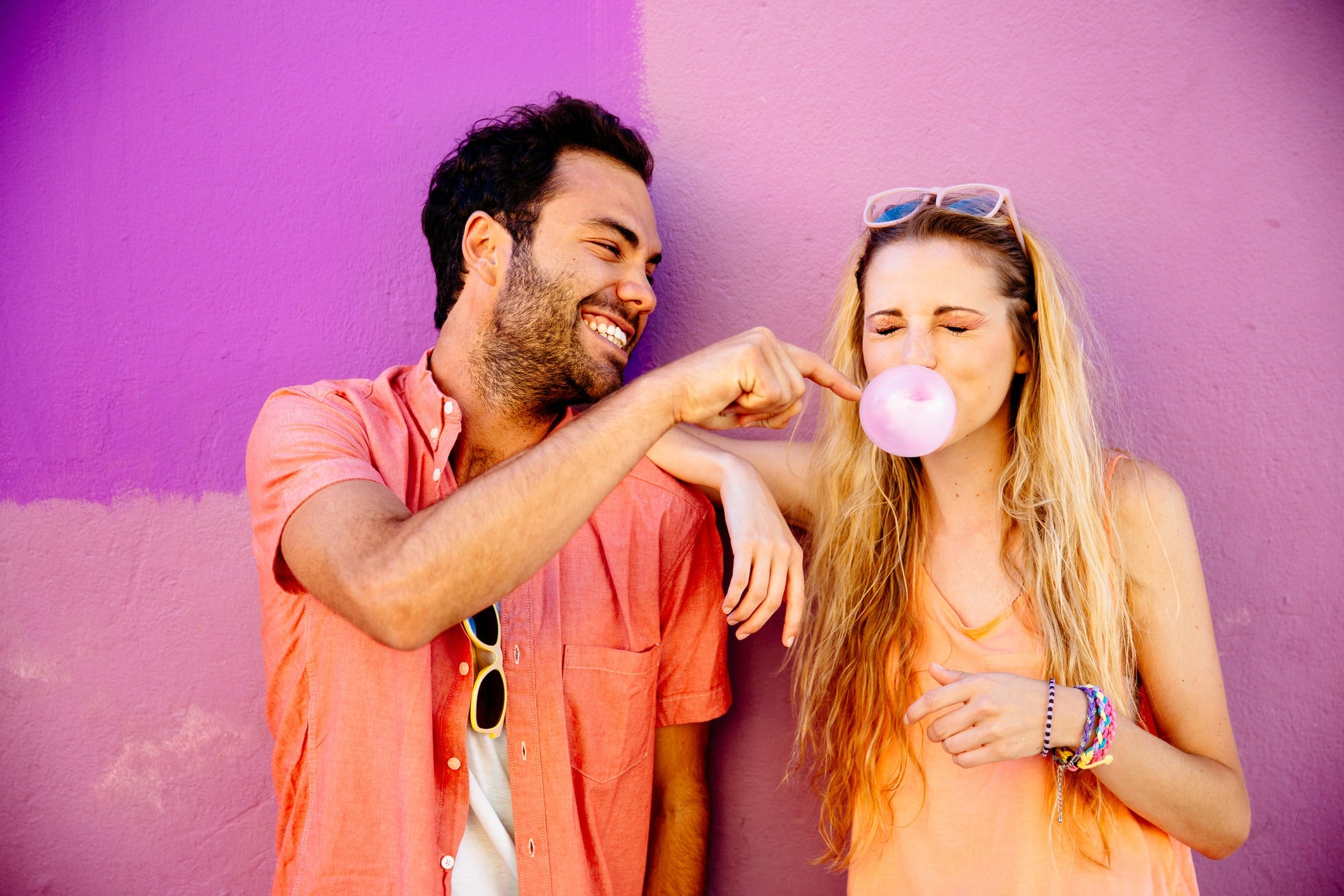 Young couple against pink background. Woman is blowing a bubble in bubblegum.