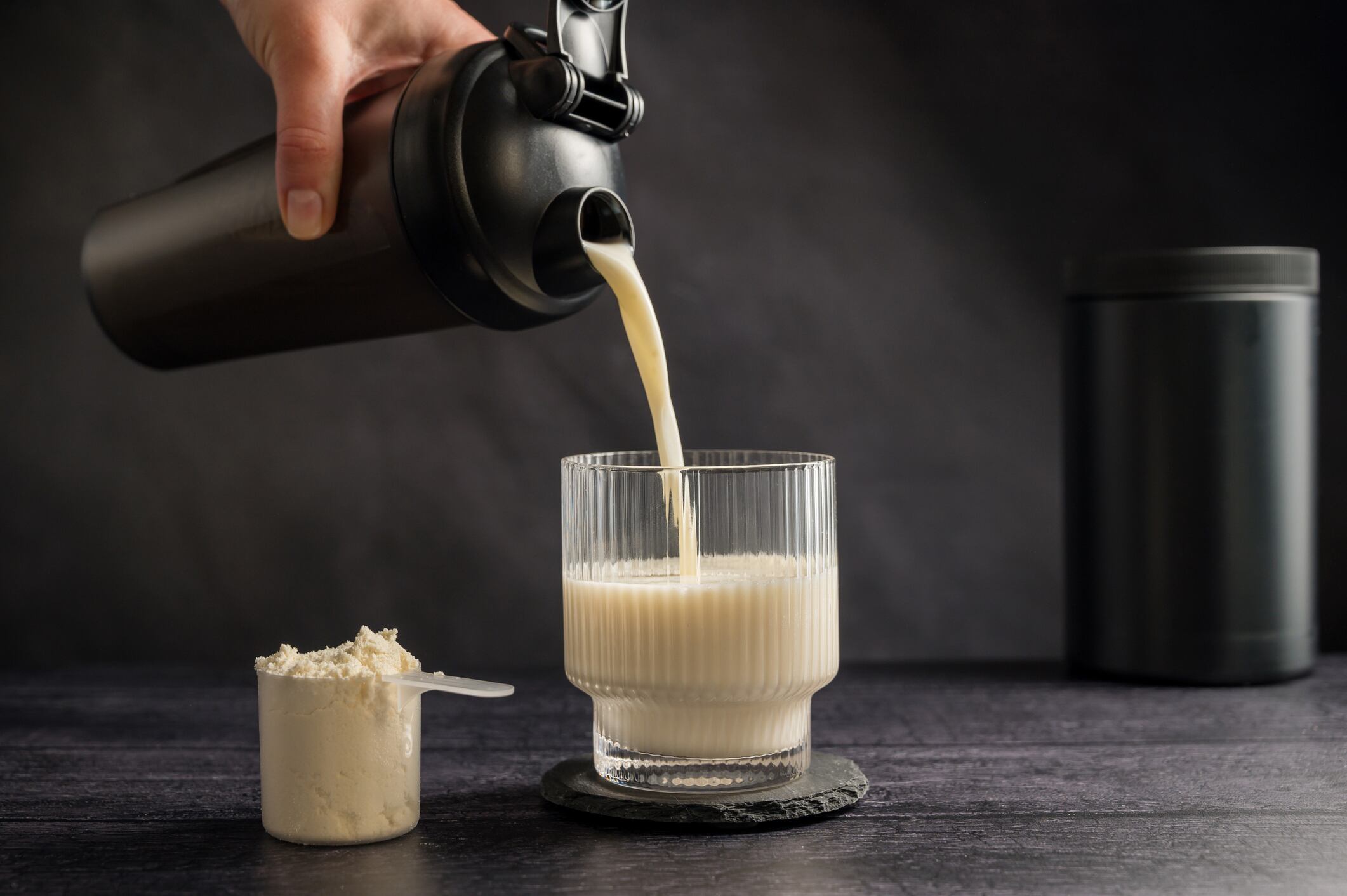 Protein drink. Pouring protein shake in a glass. Dark background. Body building, muscle growth and food supplement concept