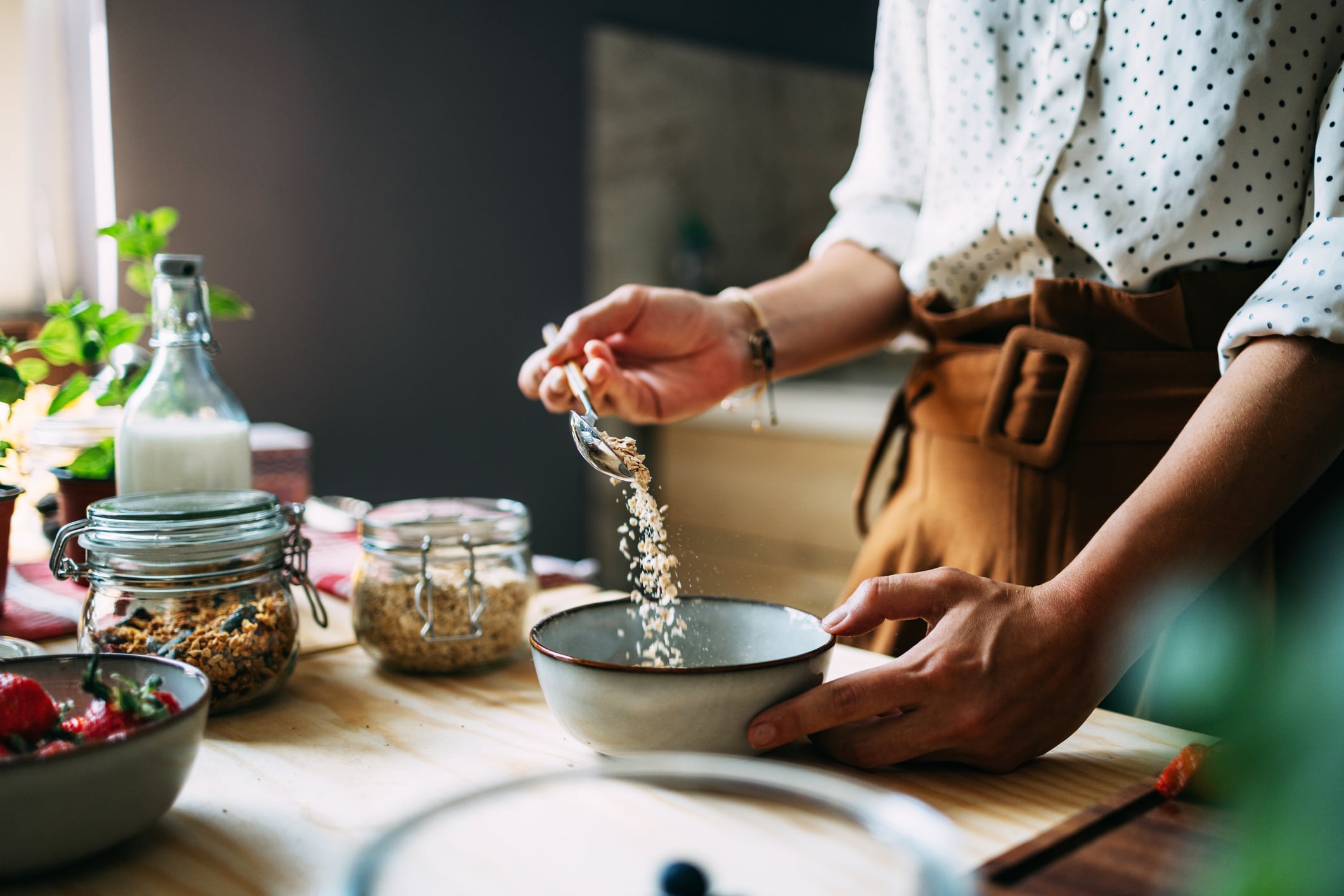 A woman in a sleeved polka dot shirt and apron prepares a healthy cereal breakfast in a sunlit kitchen, surrounded by fresh ingredients.