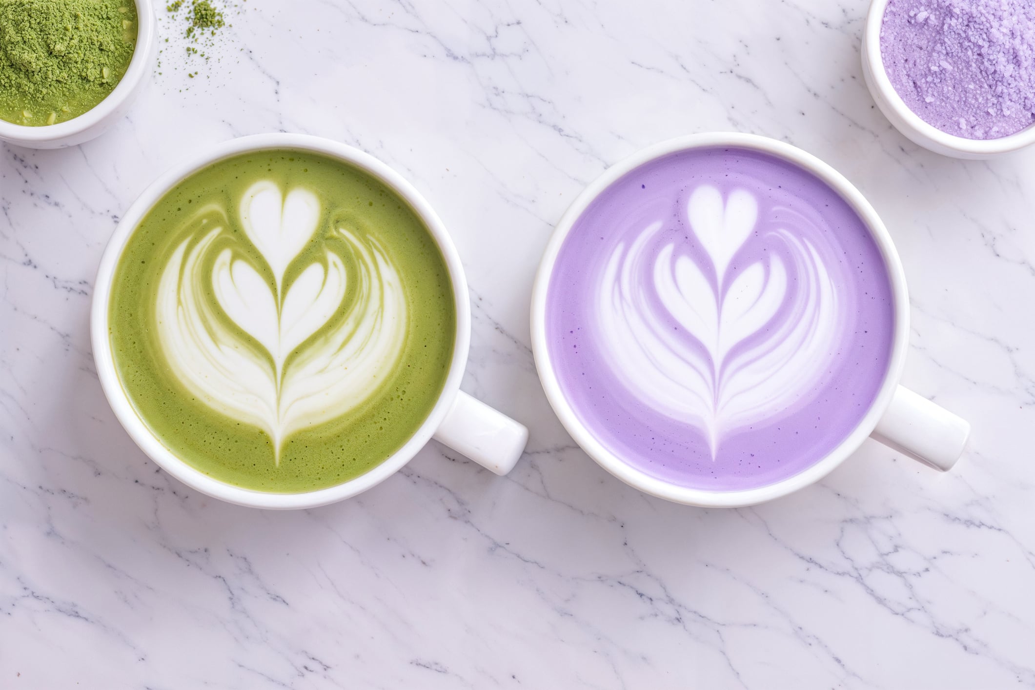 Macro top view of two colorful café beverages placed side by side: a vibrant ube latte and a green matcha latte, both topped with delicate latte art. This visually striking comparison highlights the contrast between the soft purple tones of ube purple yam and the rich green color of matcha tea. Both ingredients are widely appreciated in modern cafés for their natural origin, antioxidant properties, and association with wellness-focused lifestyles. The smooth foam textures and symmetrical composition create an elegant presentation ideal for contemporary coffee culture. This aesthetic beverage scene is perfect for café menus, specialty drink promotions, food photography, and social media content.