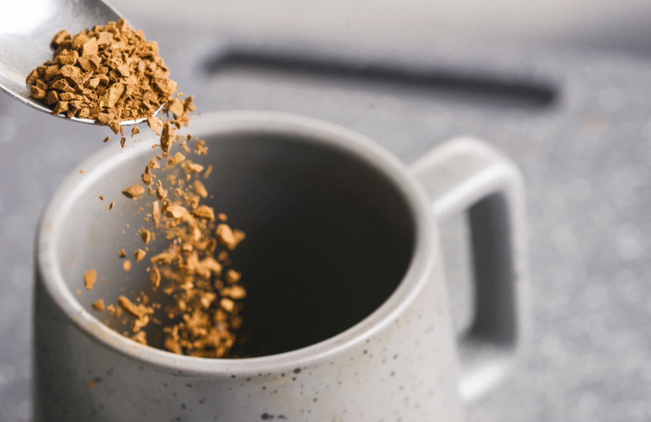 Detailed close-up of the individual grains of coffee,being tipped into a grey coffee mug,from a kitchen spoon,the movement sharply frozen,before adding boiled water.