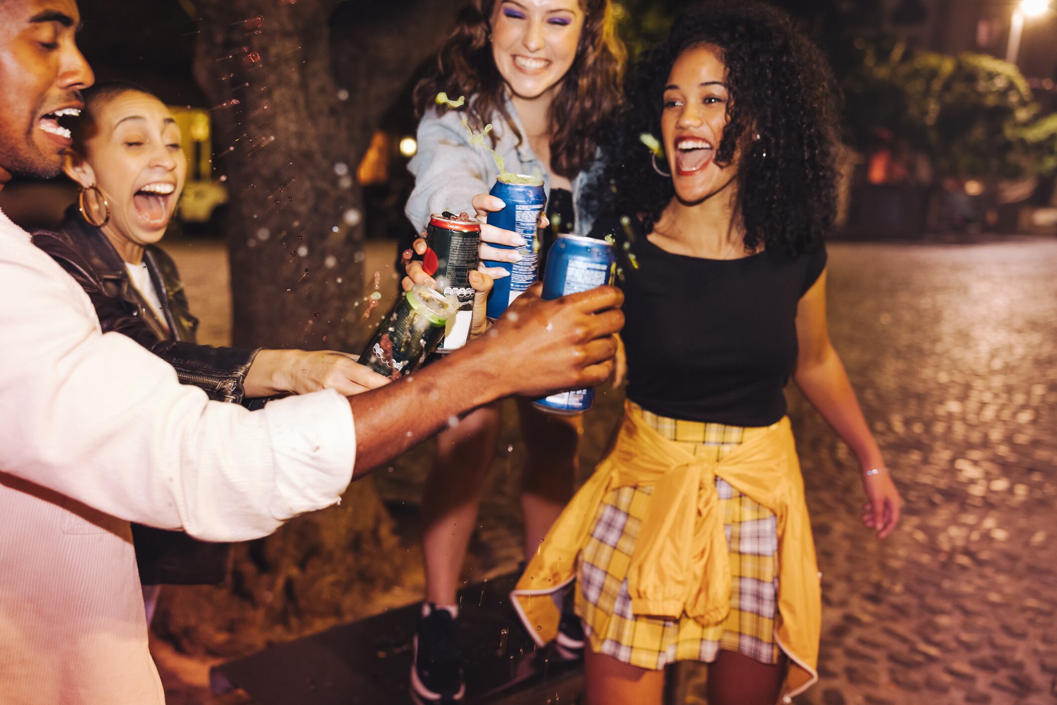 Vibrant friends toasting with spilling beer cans while standing outdoors at night. Group of ecstatic young people celebrating new year's eve with cold drinks.