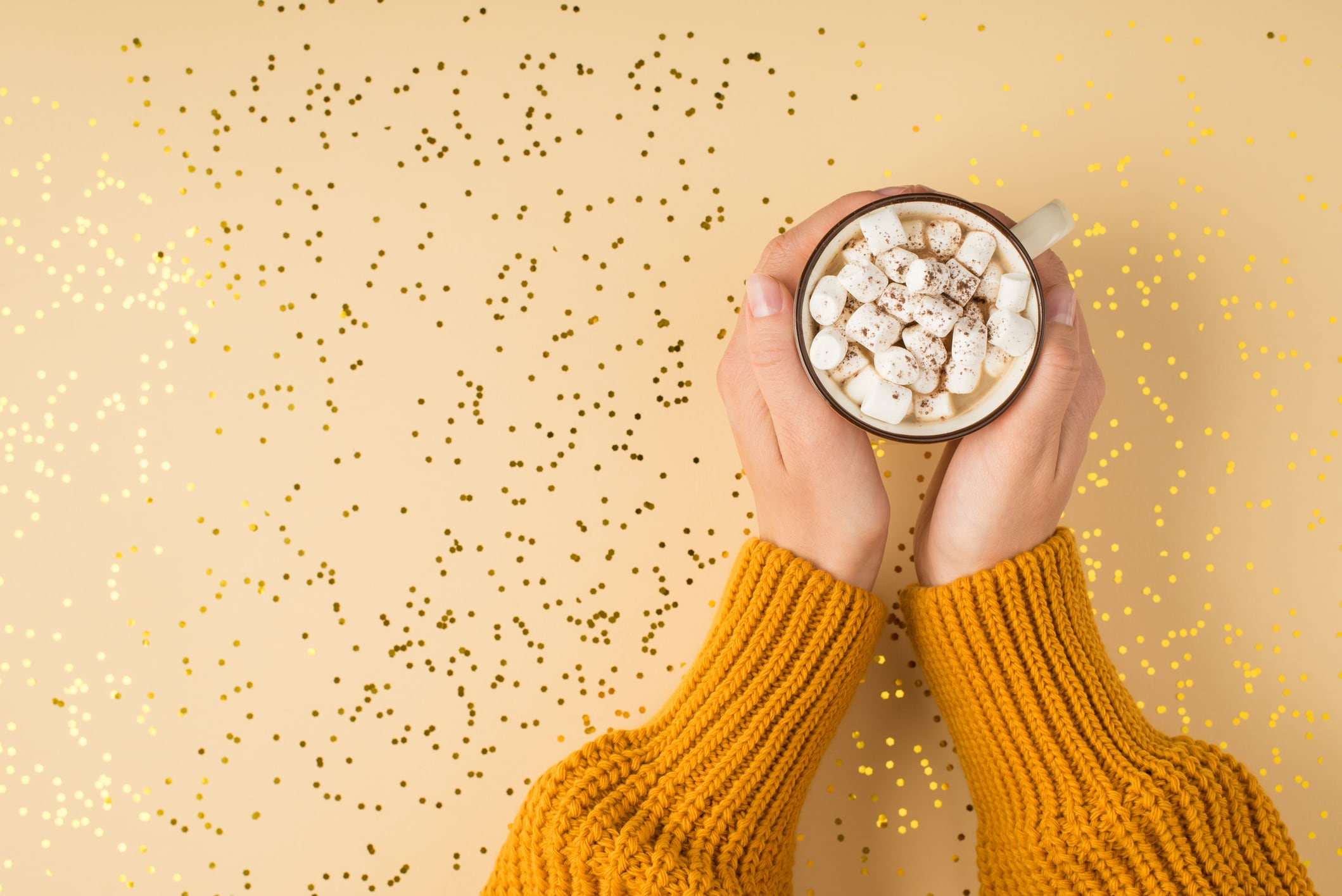 First person top view photo of female hands in yellow pullover holding mug of drink with marshmallow over shiny golden sequins on isolated pastel orange background with copyspace