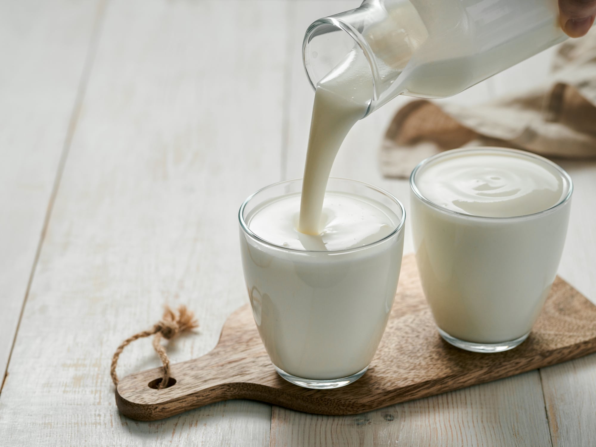 Pouring homemade kefir, buttermilk or yogurt with probiotics. Yogurt flowing from glass bottle on white wooden background. Probiotic cold fermented dairy drink. Trendy food and drink. Copy space left