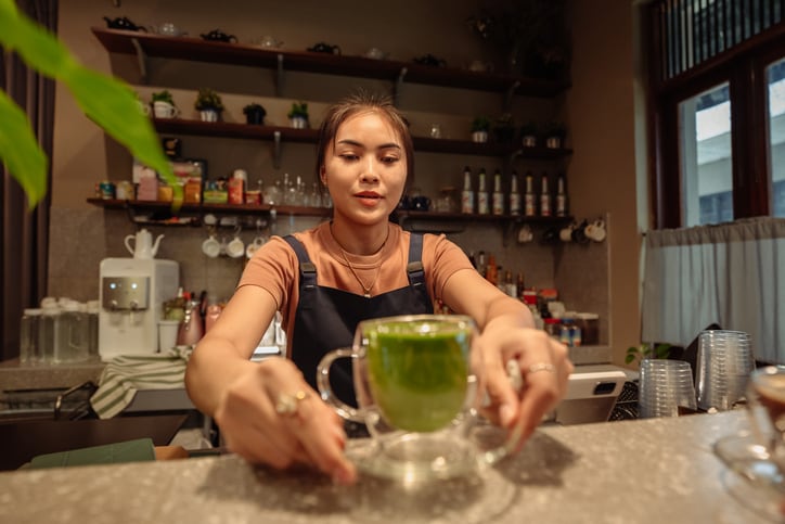 A female barista placing a glass cup of matcha latte on the café counter.