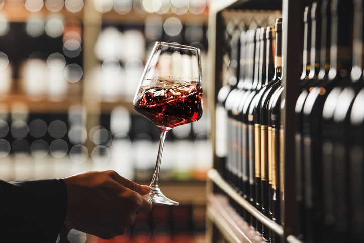 Close up shot of person hand as they swirl a glass of red wine, demonstrating the technique used to aerate the wine and release its aromas and exploring the depth of the wines flavor profile on blurred wine cellar background.