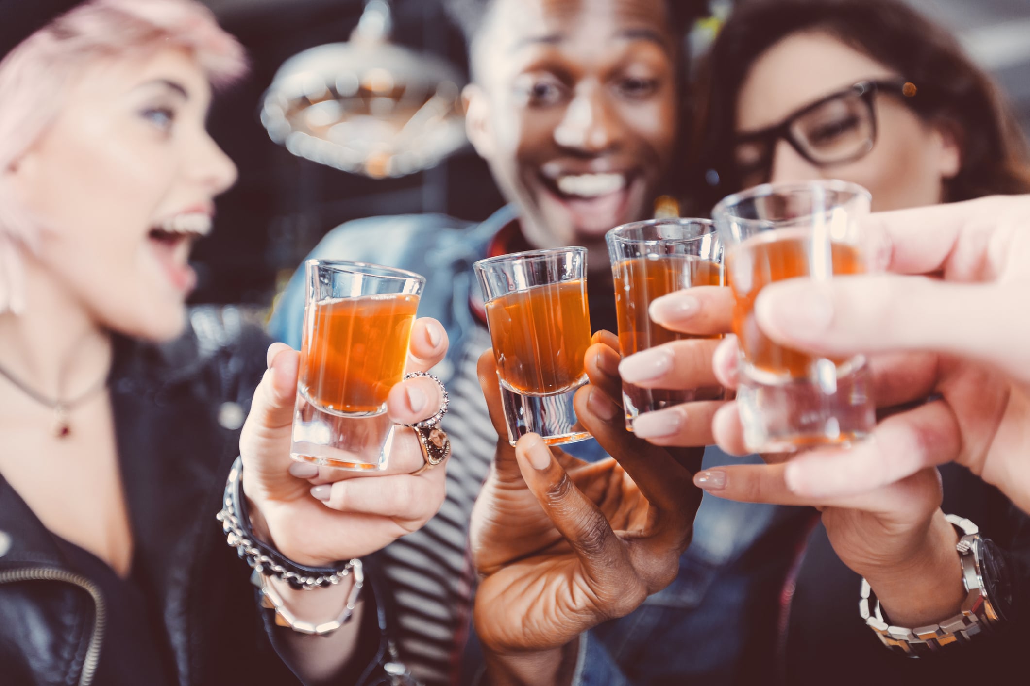 Multi ethnic group of happy friends - caucasian and afro american - drinking shots in the pub. Focus on hands and shot glasses.